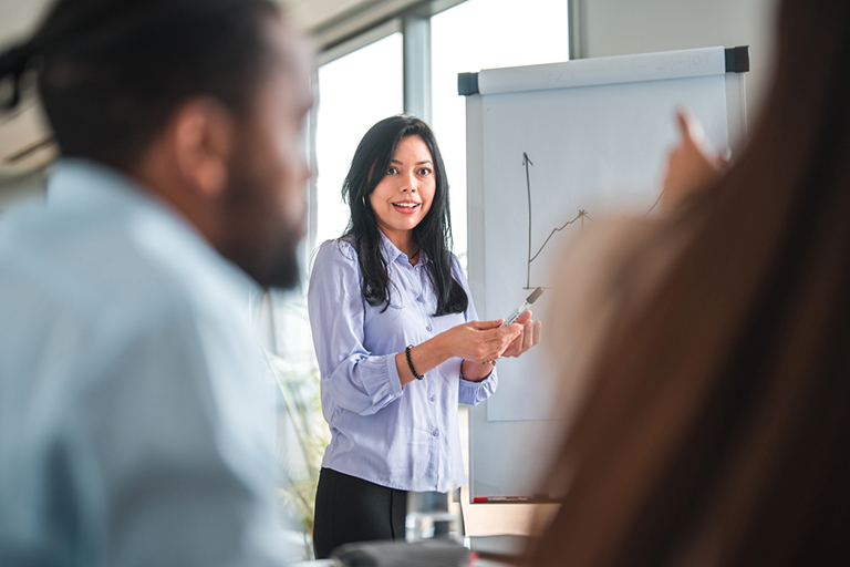 Workforce, 3/4 length image of a beautiful, smiling Latina financial expert standing as she shows a business diagram on white board. Shot over the shoulder of an African man and head of a woman in blurred foreground.