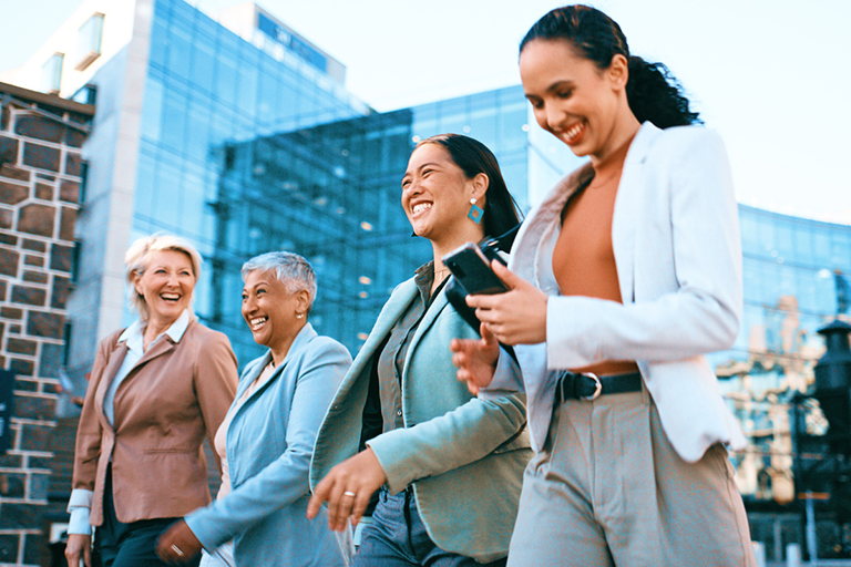 Workforce, Diverse group of professional women walking, smiling, and using a smartphone outdoors in a modern business district