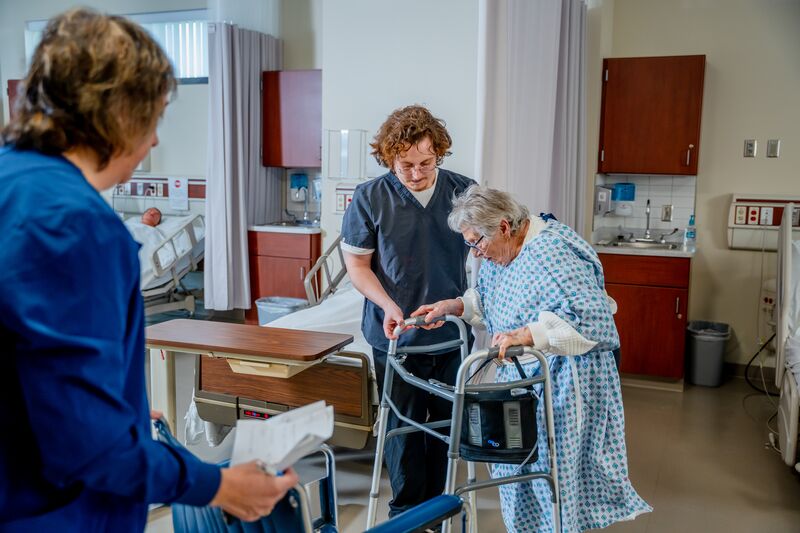 certified nurse aide (CNA), lab, male student, instructor and female patient walking with walker