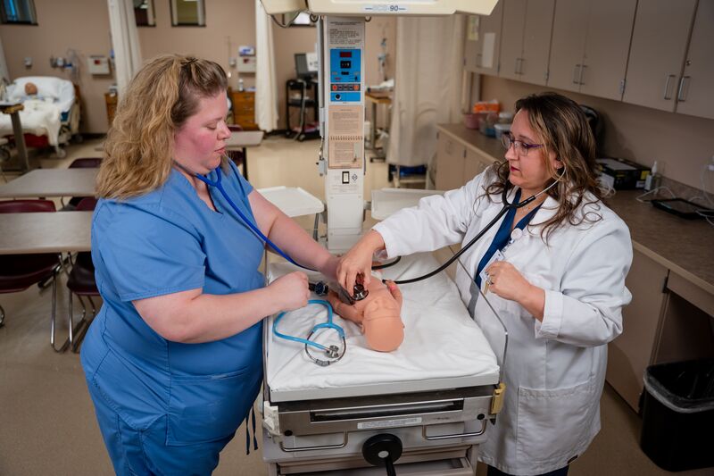 RN completion for LPN, female student listening to heartbeat on infant nursing simulator with instructor
