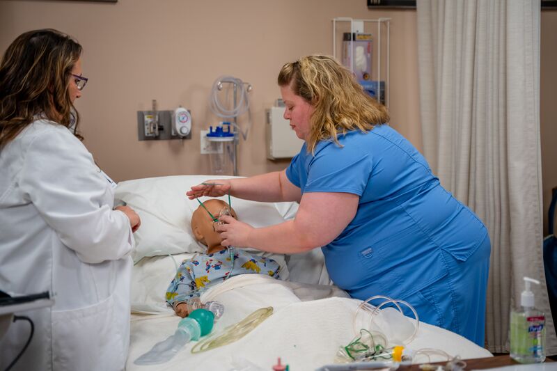 RN completion for LPN, female student adding oxygen mask to infant simulator as instructor watches