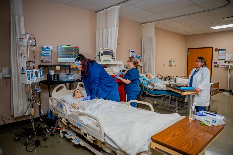 RN completion for LPN, two female students perform CPR to nursing simulator as instructor watches and grades them