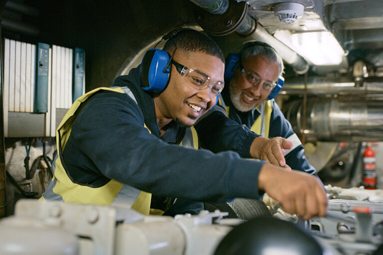 Workforce, Senior expert mechanic coaching a trainee in a manufacturing plant. Smiling men in uniform working on engine repair in factory wearing safety glasses. Professional mechanic in development