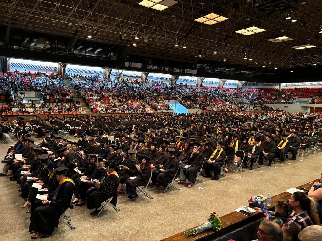 Commencement ceremony inside large civic center, hundreds of graduates seated on the floor while guests sit in the stands surrounding the graduates