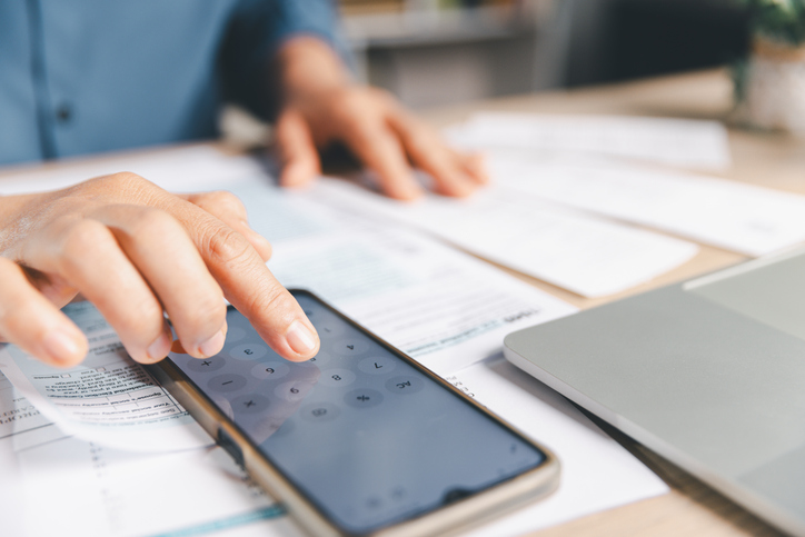 A person using a smartphone calculator app to compute finances on a desk with tax forms and a laptop. Perfect for tax preparation, accounting, business planning, and personal budget management themes.