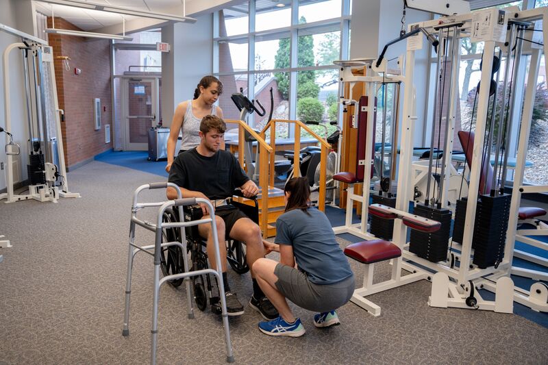 Physical therapy assisting program, two female students helping male student to stand out of wheelchair