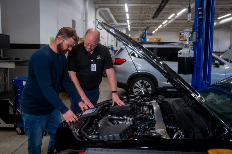 male automotive student receiving instruction from male instructor, under hood of car