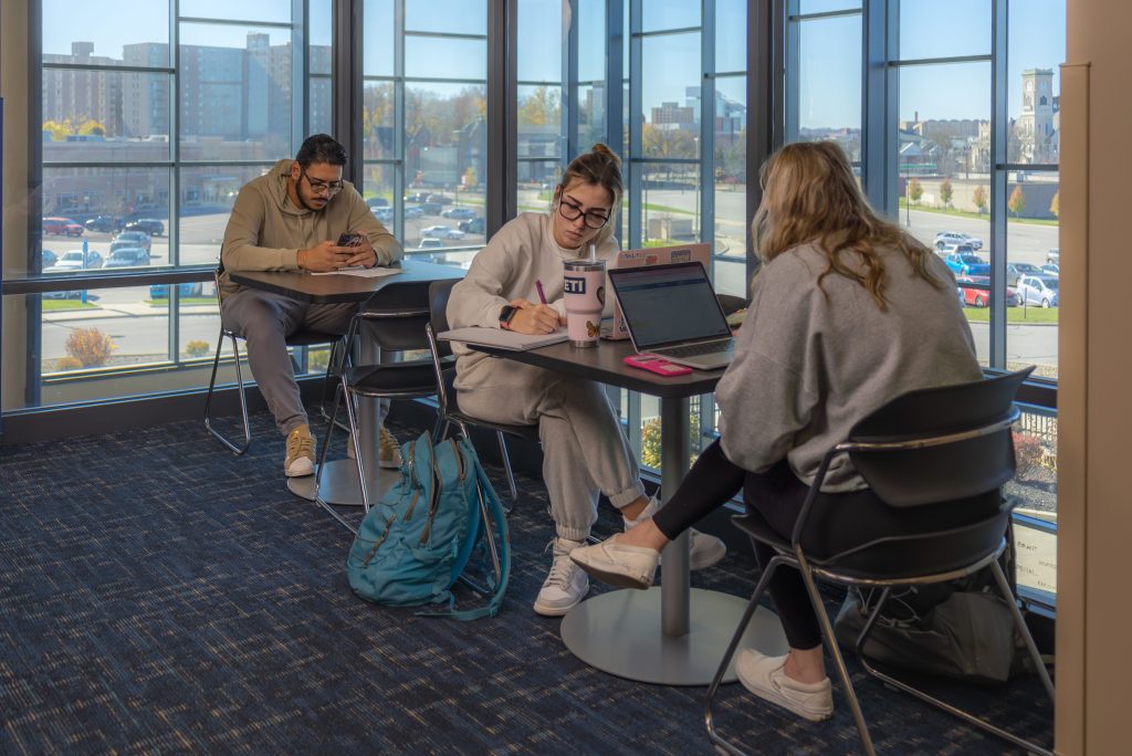 Transients, three students studying at a table, one is on his phone