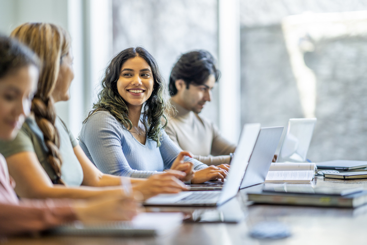 A small group of four mature adult students sit together at a long desk as they study with one another. They have text books and laptops out in front of them as they take notes and talk amongst themselves.