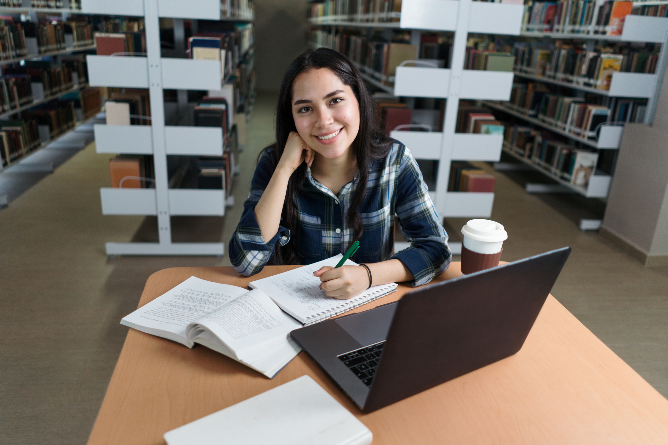 CCP, Female student doing homework at a table in the library and smiling at the camera.