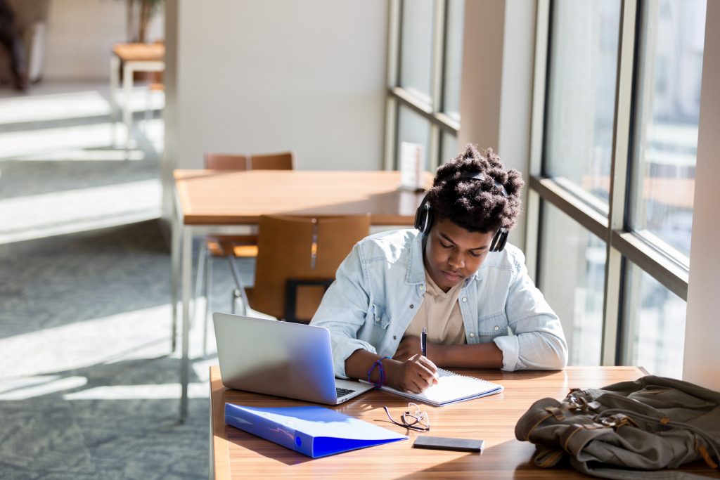 UBMS, African American teenage boy writes something in a notebook while studying in the campus library. An open laptop is on the table. He is wearing wireless headphones.