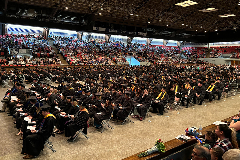 Commencement ceremony inside large civic center, hundreds of graduates seated on the floor while guests sit in the stands surrounding the graduates